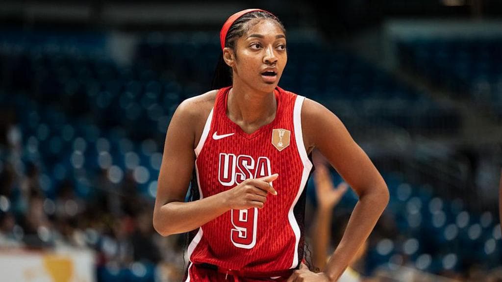 Angel Reese stands on an indoor court wearing a red USA basketball jersey with the number nine on the front and a red headband. One hand rests near the waist while the other arm is raised slightly, pointing forward. Blue arena seats and blurred figures are visible in the background under bright lighting