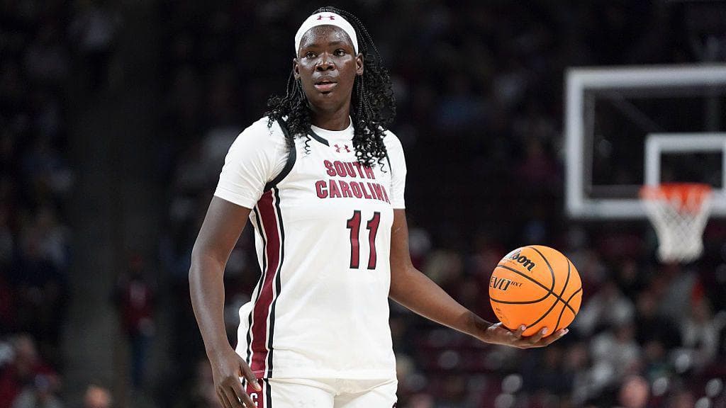 Basketball player Madina Okot stands on an indoor court holding an orange basketball at waist height in her left hand. She wears a white South Carolina jersey with the number 11 on the chest and a white headband, with long dark hair falling over the shoulders. A basketball hoop and blurred spectators are visible in the background under bright arena lighting