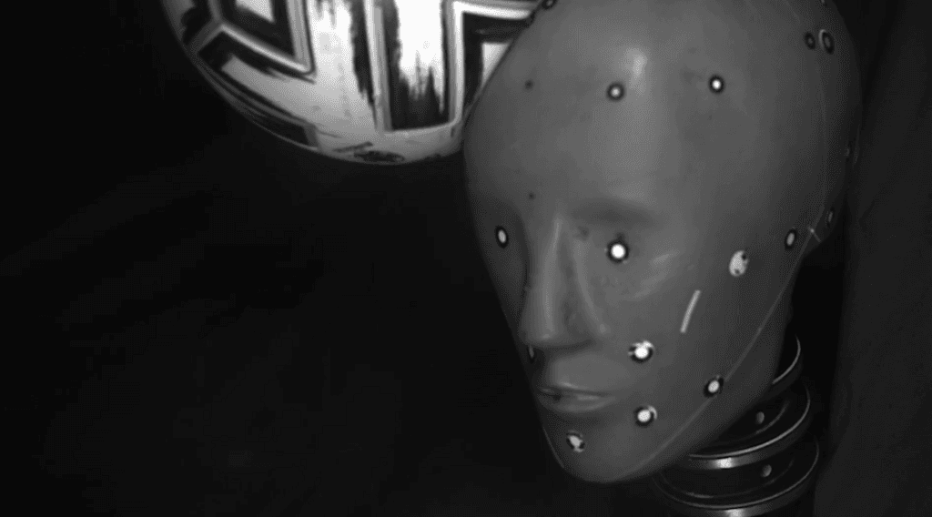The grey head of a crash dummy, with white sensors at various points on the face, being hit by a white and black football on its right-hand side against a black background