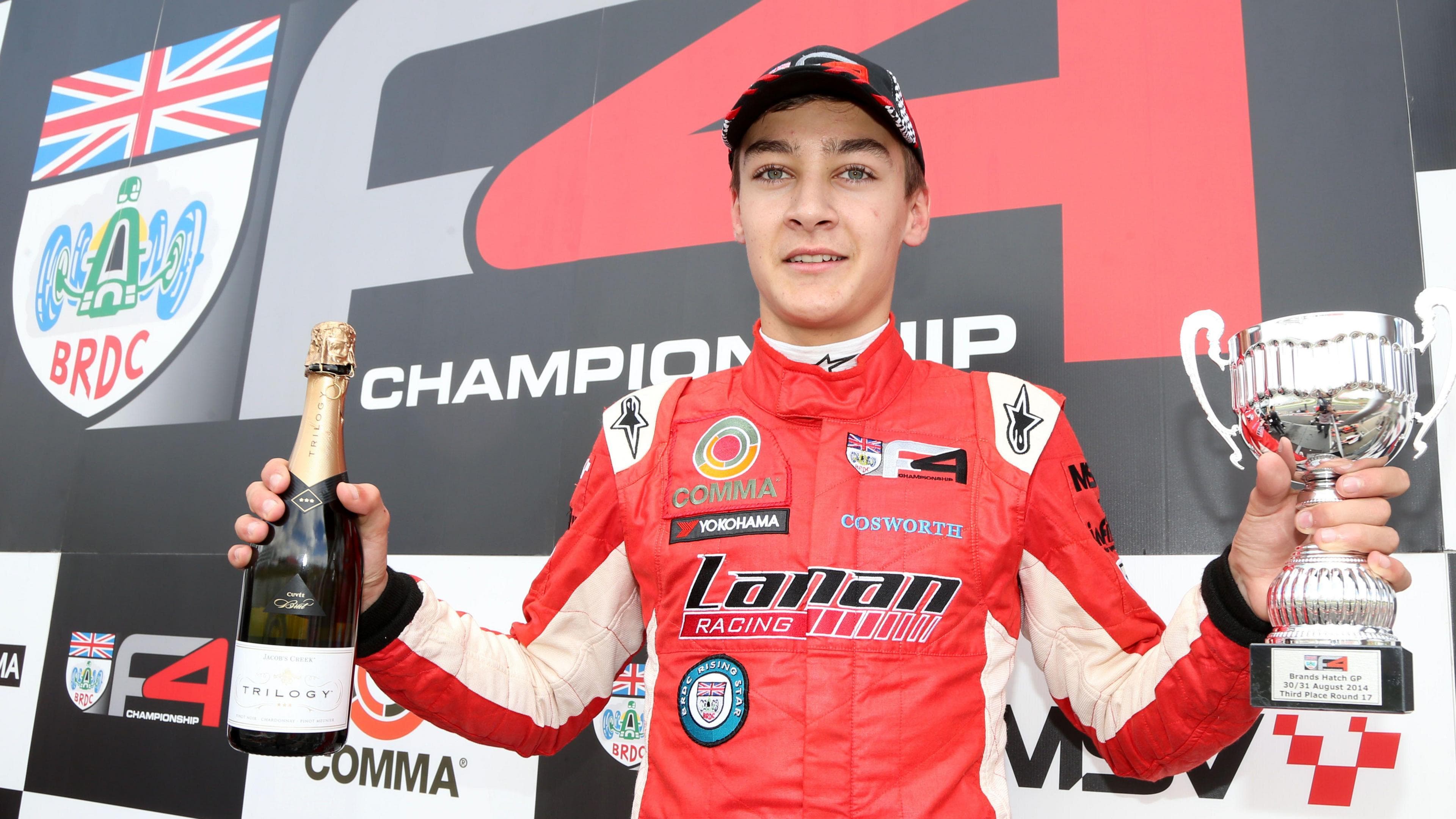 George Russell celebrates on a podium after a Formula 4 victory at Brands Hatch in 2014. He is wearing a red racing suit and his holding a small glass bottle of a drink in his right hand and a trophy in his left hand