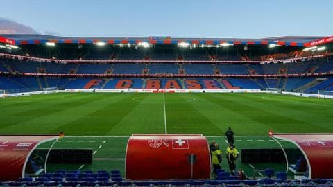 View of St Jakob-Park stadium taken from behind a red dugout on the halfway line, with the name FC Basel in red text on a blue background on the seats opposite