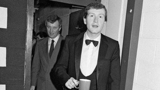 Steve Davis, in dark dinner suit and bowtie, holds a coffee cup and looks towards the camera backstage at the Crucible Theatre, as a suited Barry Hearn stands just behind him